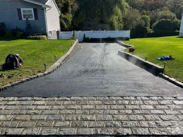 Newly paved black asphalt driveway with brick border and house in background.
