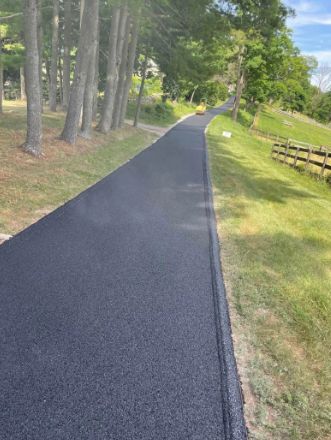 Newly paved asphalt driveway with trees on the left and grassy hill with fence on the right.