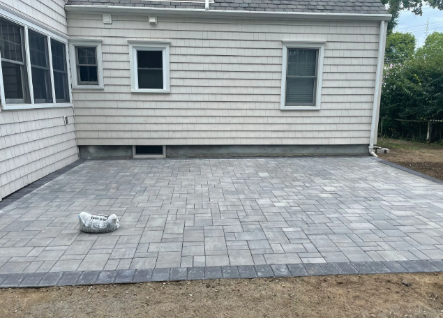 Gray paver patio next to a light-colored house with three windows.