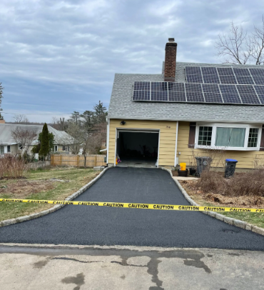 Freshly paved black driveway in front of a yellow house with solar panels; caution tape across.