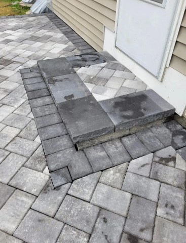 Gray paver patio with a step leading to a white door; corner of a beige house.