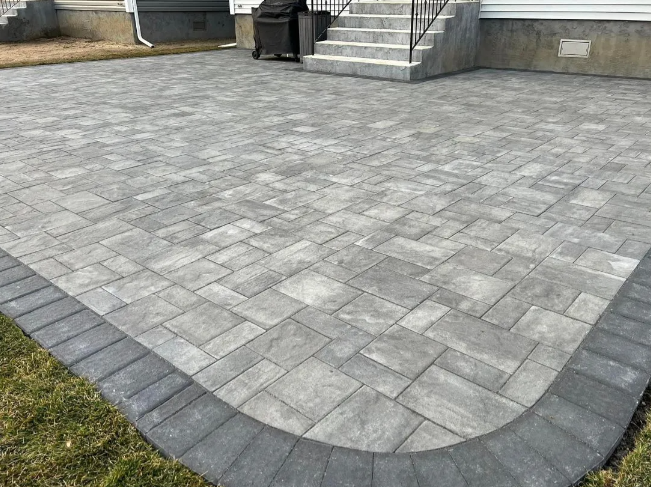 Gray paver patio with a dark border next to green grass and stairs.