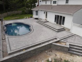Backyard patio with pool, brickwork, and raised platform with steps, alongside a white house with glass doors.