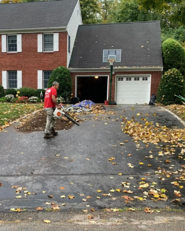 A person in red shirt uses a leaf blower to clear leaves from a driveway in front of a house.