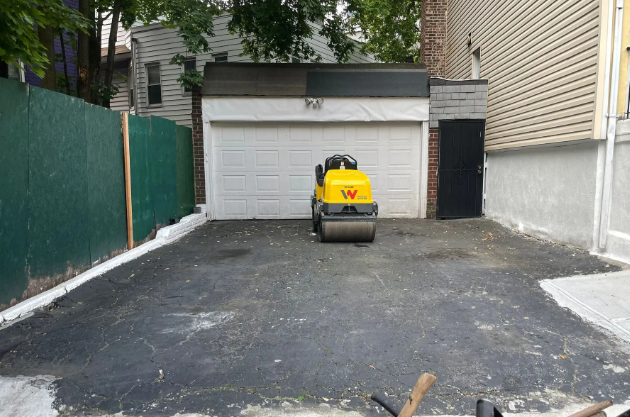 Asphalt driveway with roller, garage, and green fence.