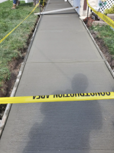 Freshly poured concrete sidewalk with yellow caution tape, bordered by grass and a house.