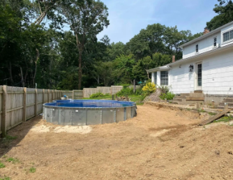 Backyard with an above-ground pool and a white house. Sand area around the pool, wooden fence, and trees.