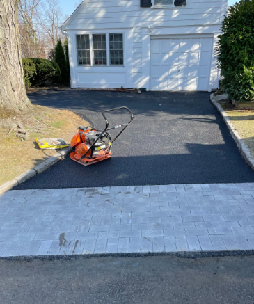 Asphalt driveway being installed next to a gray paver walkway, with a compactor machine on the new asphalt.