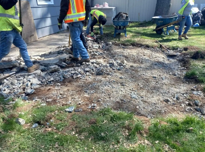 Construction workers in vests removing debris from an outdoor area with tools and a wheelbarrow.
