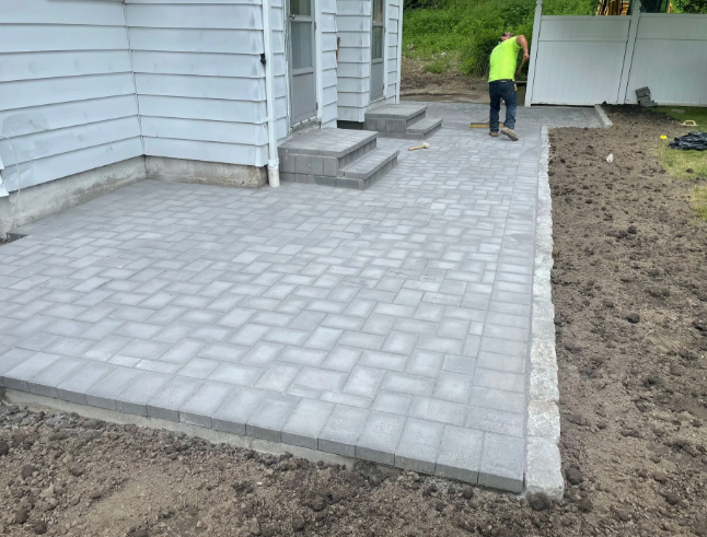 Brick patio being installed by a worker; near a house with steps.