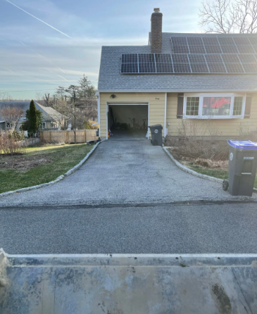 Asphalt driveway leading to a yellow house with solar panels, garage, and trash bins.