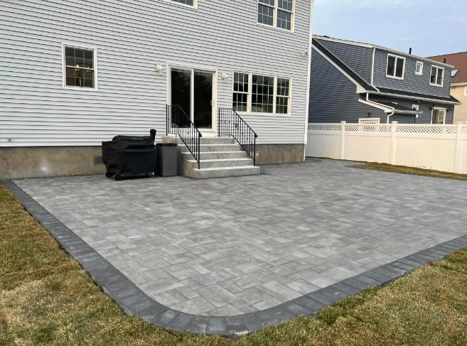 Backyard patio with gray pavers and a bordering of darker pavers, adjacent to a light gray house.