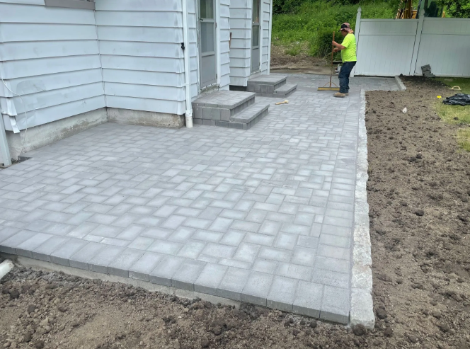 Grey brick patio next to a house with steps. A person in a yellow shirt works nearby.