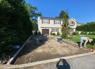 Driveway construction at a two-story house. A worker is near the driveway edge, tools and wheelbarrows are visible.