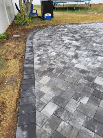 Brick patio with black border, next to a grassy area, with a fence and tree in the background.
