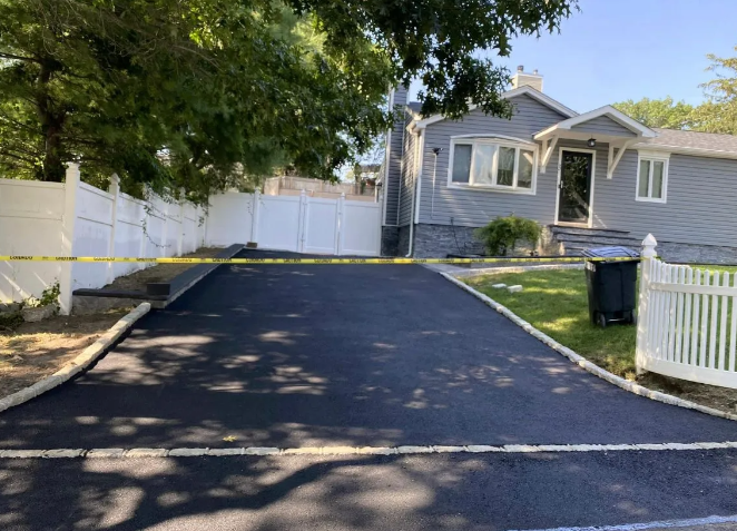 Newly paved asphalt driveway leading to a gray house, bordered by white fences and a green lawn.