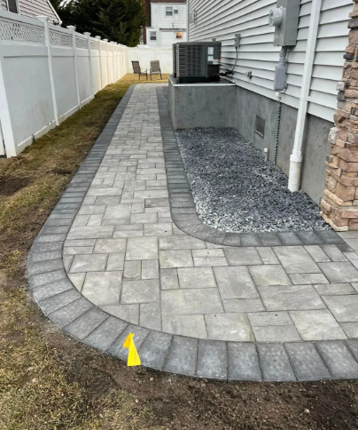 Brick pathway curves around a house, bordered by grass and a white fence.