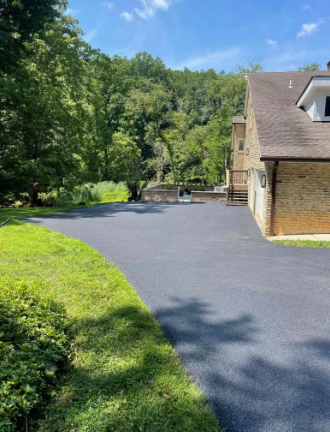 Newly paved asphalt driveway next to a brick house and grassy area with a wooded hillside in the background.