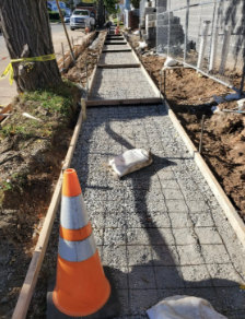 Sidewalk construction: gravel and rebar framework in wooden forms, orange traffic cone in foreground.
