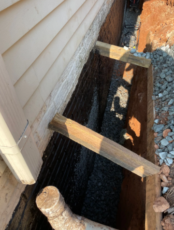 Construction site: foundation trench with gravel, wire mesh, wooden supports, and house siding.