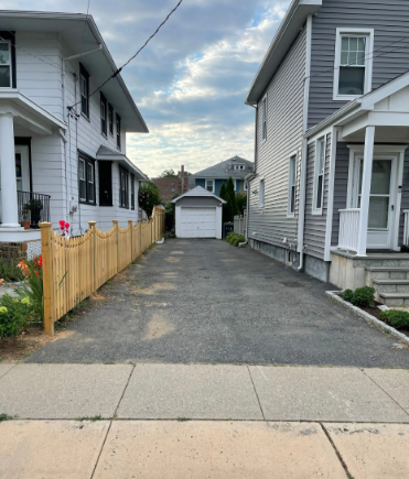 Narrow paved driveway between two houses, leading to a detached garage. Wooden fence on left, sidewalk in foreground.