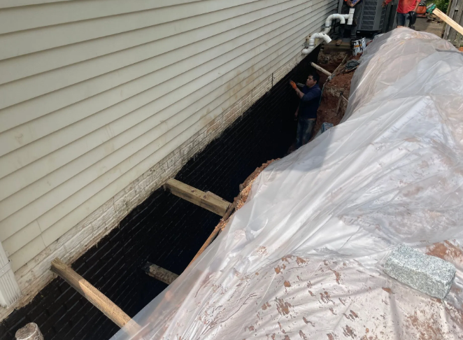 Construction: foundation waterproofing, house side view, trench, black membrane, worker, plastic tarp, soil, white siding.