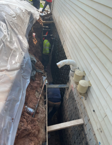 Workers excavating next to a building's white siding. One person is in the trench, installing a drainage layer.
