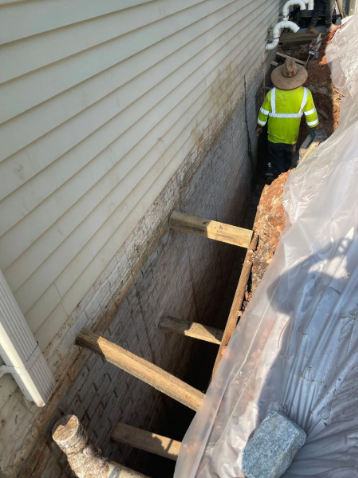 Worker in high-vis vest stands in a trench next to a house foundation with wooden supports.