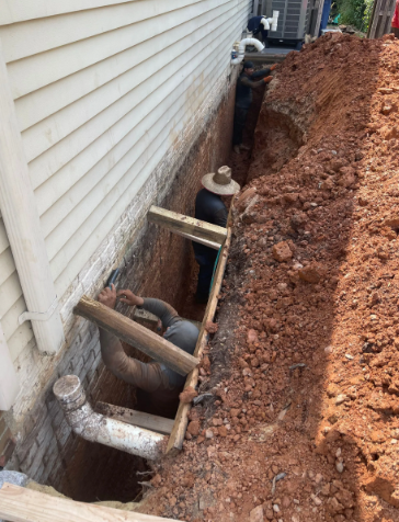 Workers in a trench alongside a house, repairing pipes. One wears a hat. Earth-toned colors.