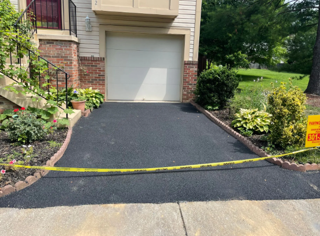 Freshly paved driveway with yellow caution tape, bordered by landscaping and a garage.