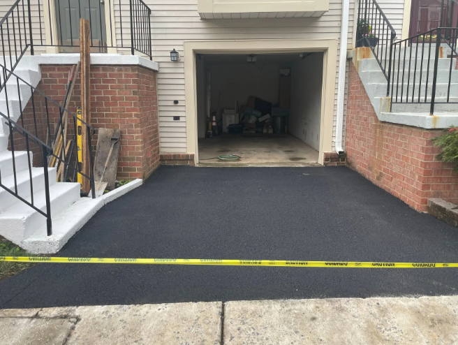 Freshly paved black asphalt driveway in front of a tan garage and two-story townhome. Caution tape present.
