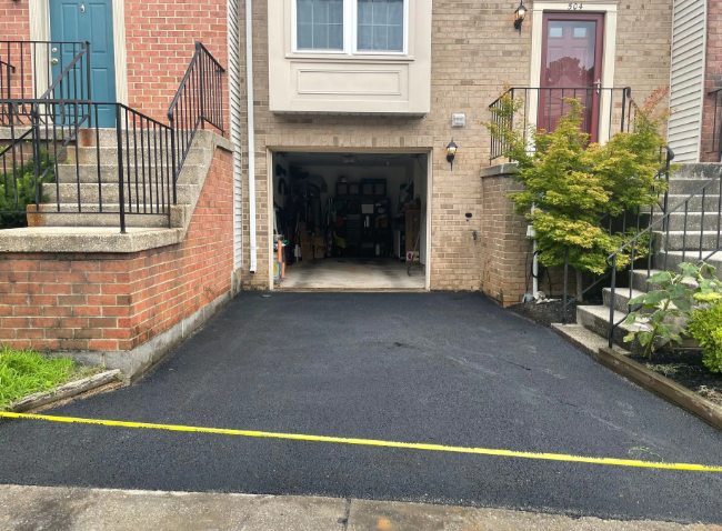 Asphalt driveway in front of a brick townhome with an open garage and steps leading up to the entrance.