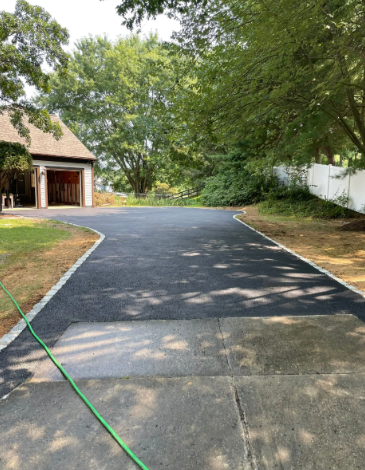 Asphalt driveway with concrete border, leading to a garage. Green grass and trees frame the scene.