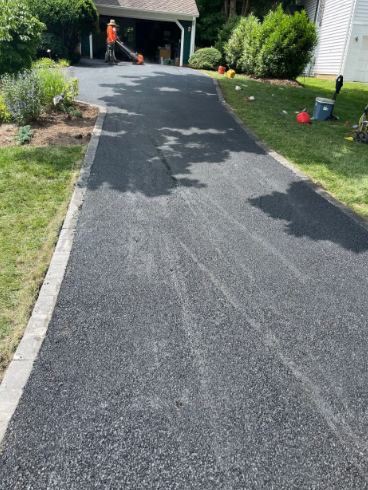 Newly paved driveway with a person operating a leaf blower in the background. Green grass borders the driveway.