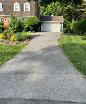Asphalt driveway leading to a white garage door in front of a house, surrounded by lawn and landscaping.