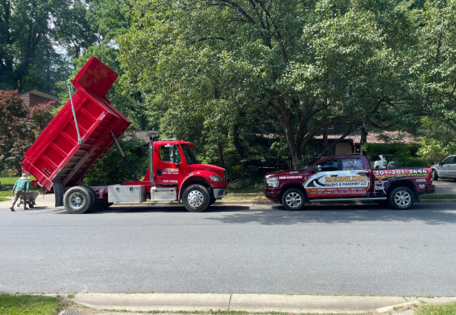 Red dump truck with raised bed next to a red pickup truck parked on a street. Man in green shirt pushing a wheelbarrow.