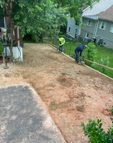 Workers preparing a dirt driveway for paving, with tools and wooden borders. Houses and trees in the background.