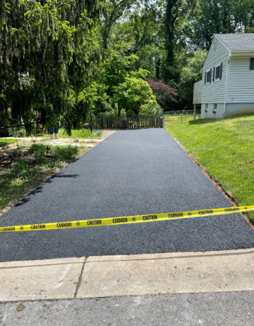 Newly paved asphalt driveway with caution tape, flanked by grass and a house.