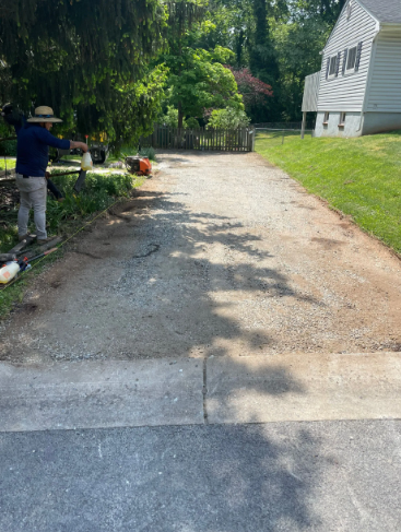Person using a leaf blower on a gravel driveway next to a house with a green lawn.