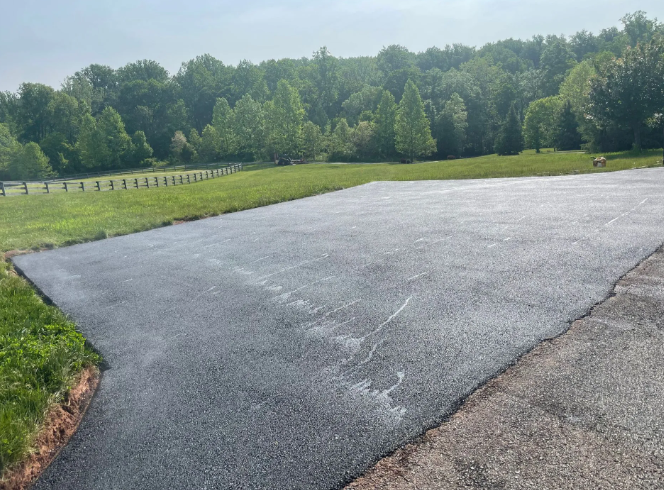 A gravel parking area transitions to grassy field. A wooden fence and trees are visible in the background.