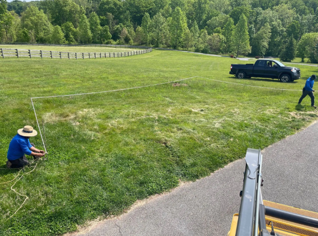 Two workers measure a rectangle in a grassy field. A black truck and a wooded fence are in the background.