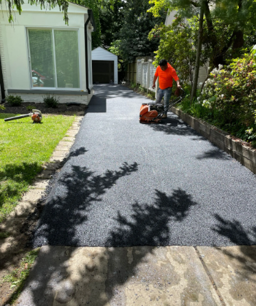 Man operating a compactor on a newly paved asphalt driveway. A white building and garage are in the background.