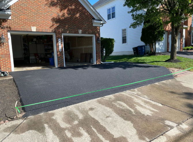 Freshly paved asphalt driveway in front of a brick garage; a green string line marks the edge.