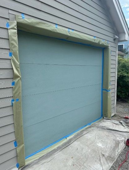 Garage door being painted, taped off with blue painter's tape and paper. The door is light blue; siding is gray.