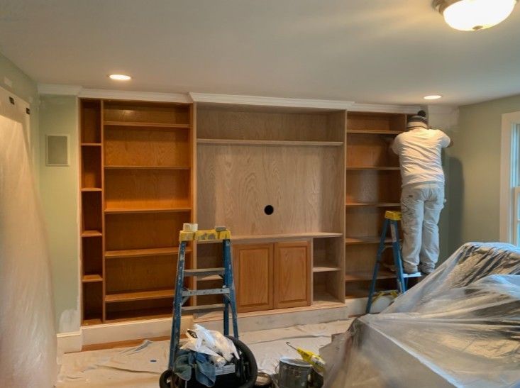Person painting a built-in wooden entertainment center with shelves. Painter on a ladder; room is partially covered in plastic.