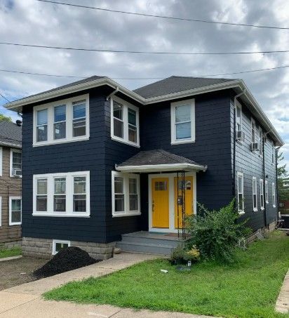Dark blue two-story house with white trim, yellow door, and grass lawn under a cloudy sky.