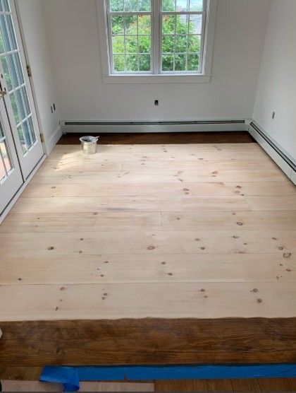 Interior room with newly installed, unfinished wood floor; doorway to left, window in the back.