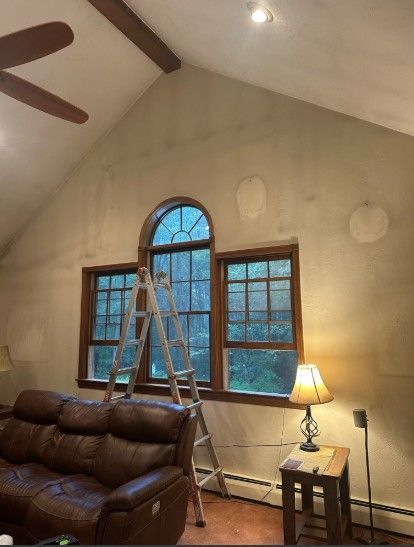 Interior room with a tall arched window, ladder, brown couch, and walls in the process of repair.