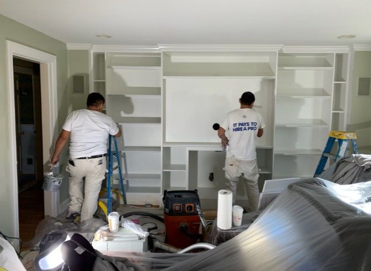 Two painters spray paint a built-in white bookshelf unit in a room.