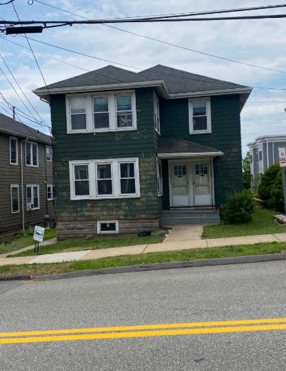 Two-story green house with peeling paint, multiple windows, and a sidewalk entrance, wires above.